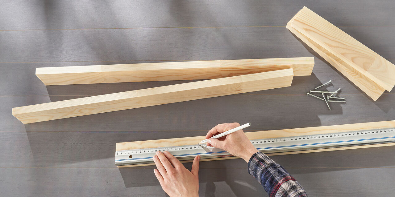 Des mains dessinent une ligne sur une planche de bois à l'aide d'un crayon et d'une règle. D'autres planches et des vis sont posées sur la table.