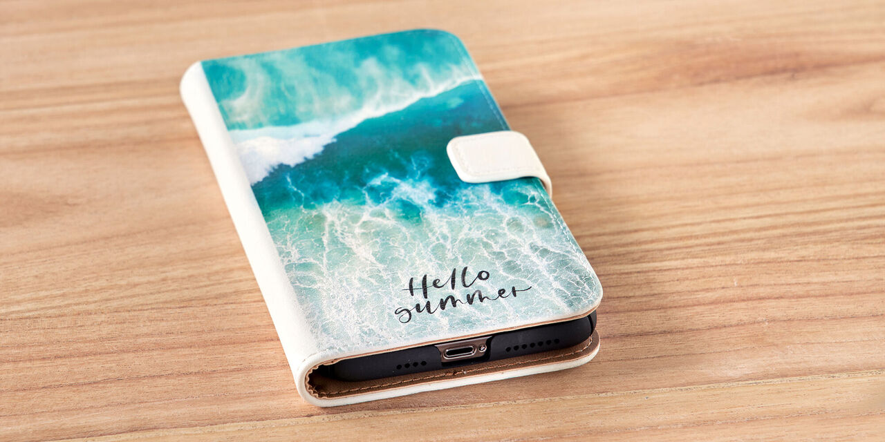 Un smartphone est posé sur une table. On y voit la coque du smartphone, sur laquelle on peut voir un gros plan d’eau et les mots « Hello Summer ». Il s’agit d’un étui sideflip en cuir blanc.