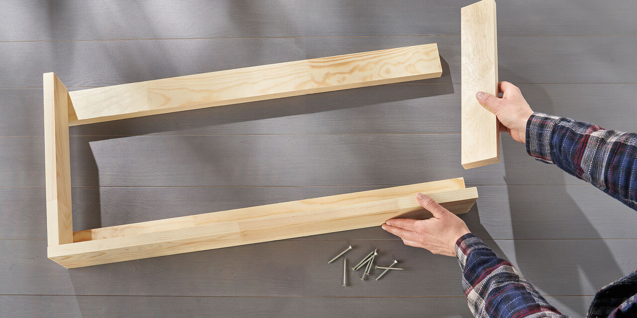 Des mains posent sur une table cinq planches de bois en forme de cadre d'étagère. Des vis sont posées à côté.